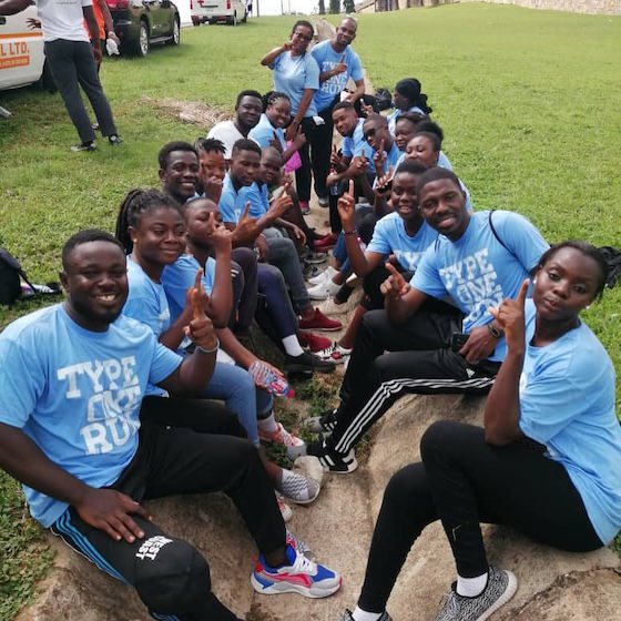 Group of young adults in light blue shirts sitting on grass and rocks, smiling and posing with peace signs