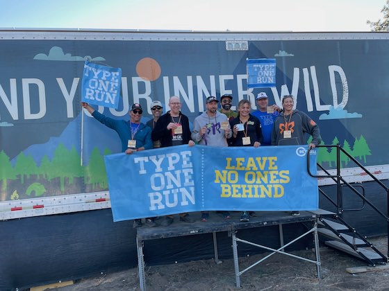Eight runners holding banners and medals posing together in front of a branded event trailer after completing a race