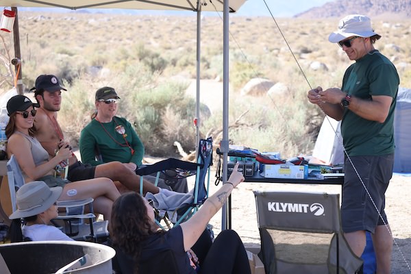 Group of five people relaxing under a canopy shade structure in a desert campsite, one person rigging a fishing rod