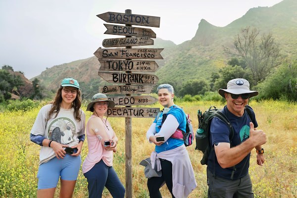Four smiling hikers with backpacks posing next to a rustic wooden directional signpost in a wildflower meadow